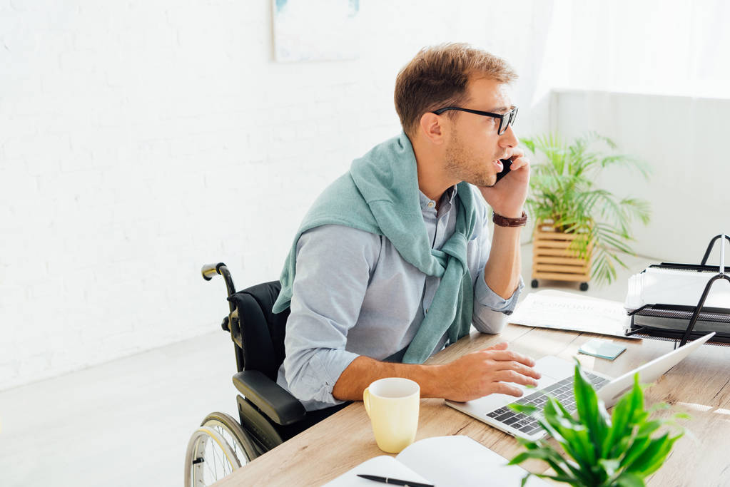 Случайный бизнесмен в инвалидной коляске разговаривает на смартфоне и использует ноутбук за столом
 - Фото, изображение