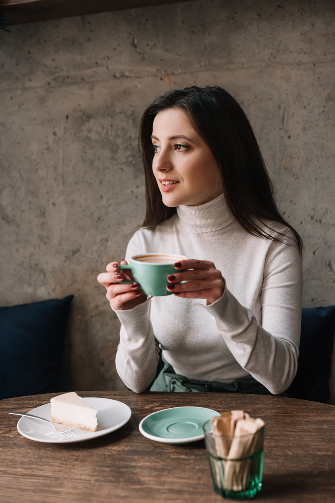 улыбающаяся женщина пьет кофе и смотрит вдаль возле чизкейка в кафе
 - Фото, изображение