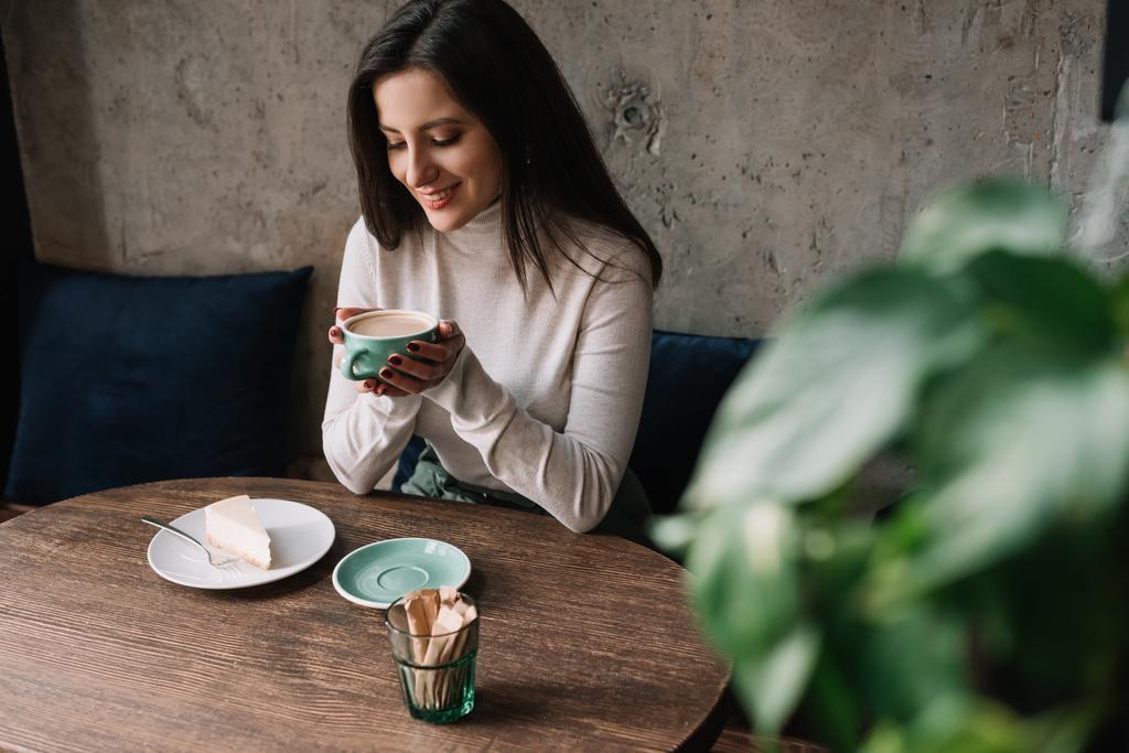 επιλεκτική εστίαση του πράσινου φυτού και χαμογελαστή γυναίκα πίνοντας καφέ κοντά cheesecake στο καφέ - Φωτογραφία, εικόνα