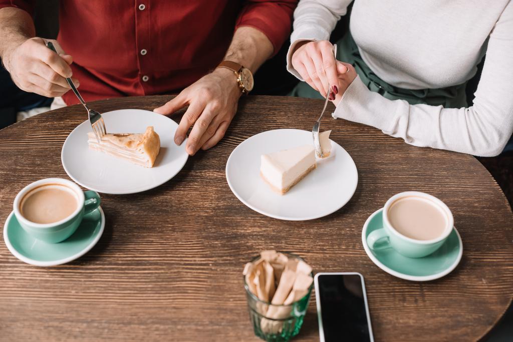kivágott kilátás pár eszik sajttorta és ivás cappuccino fából készült asztalnál okostelefon - Fotó, kép