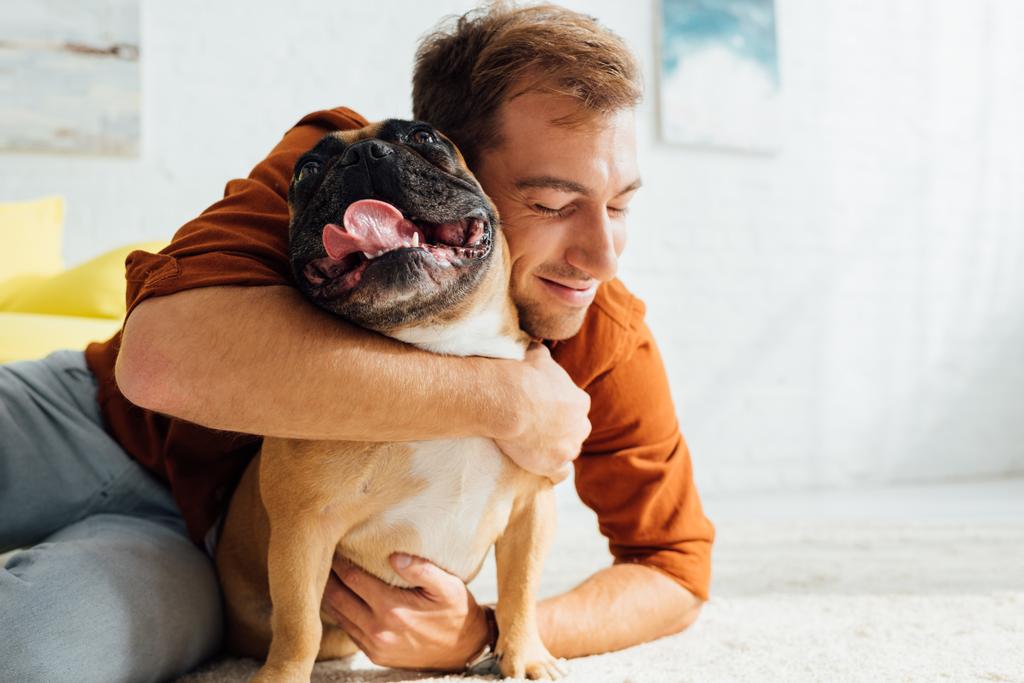 Smiling man hugging funny french bulldog on floor in living room - Photo, Image