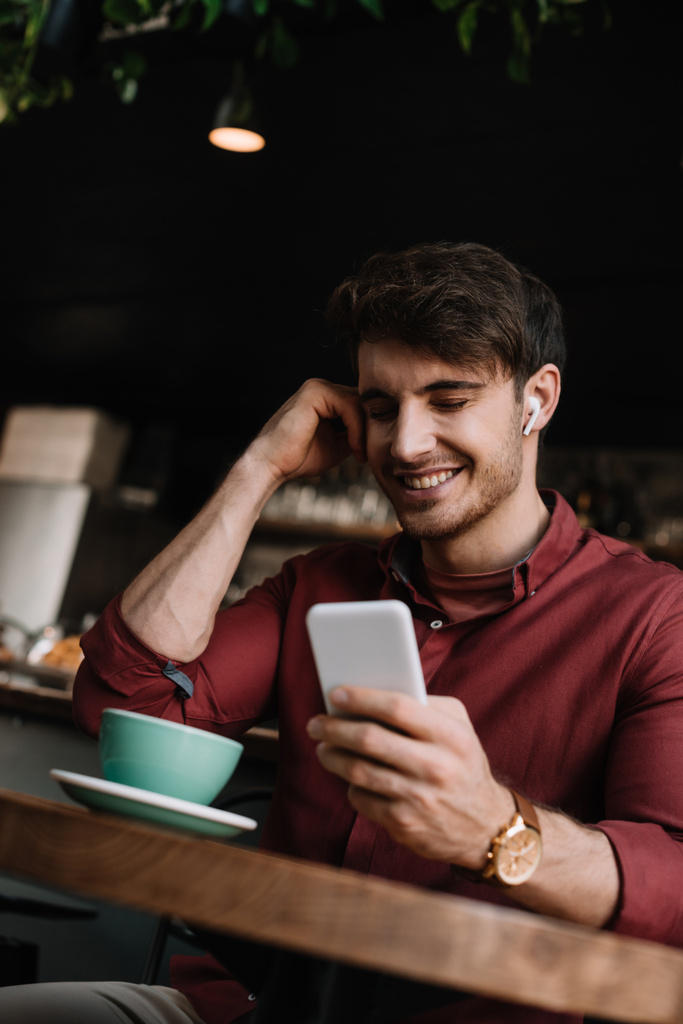 homem sorridente em fones de ouvido sem fio usando smartphone na mesa com xícara de café
 - Foto, Imagem