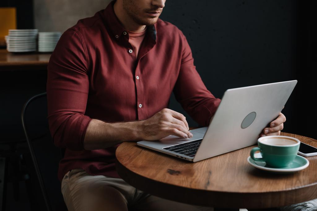 Cappuccino ve akıllı telefon yakınlarındaki kafede dizüstü bilgisayar kullanan serbest çalışan birinin görüntüsü. - Fotoğraf, Görsel