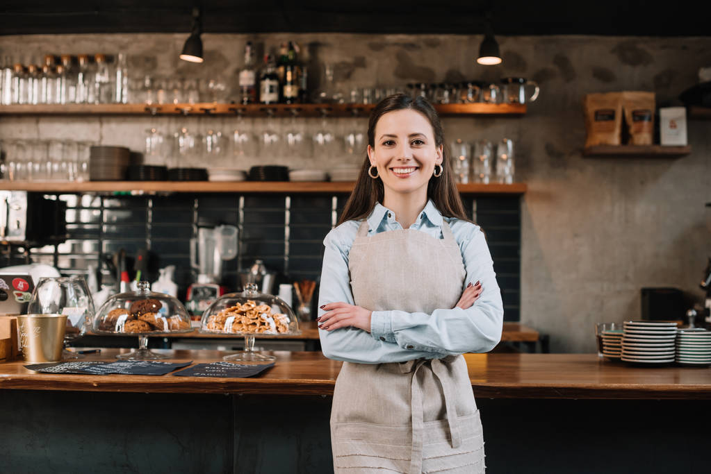barista souriant dans tablier debout avec les bras croisés près comptoir bar
 - Photo, image