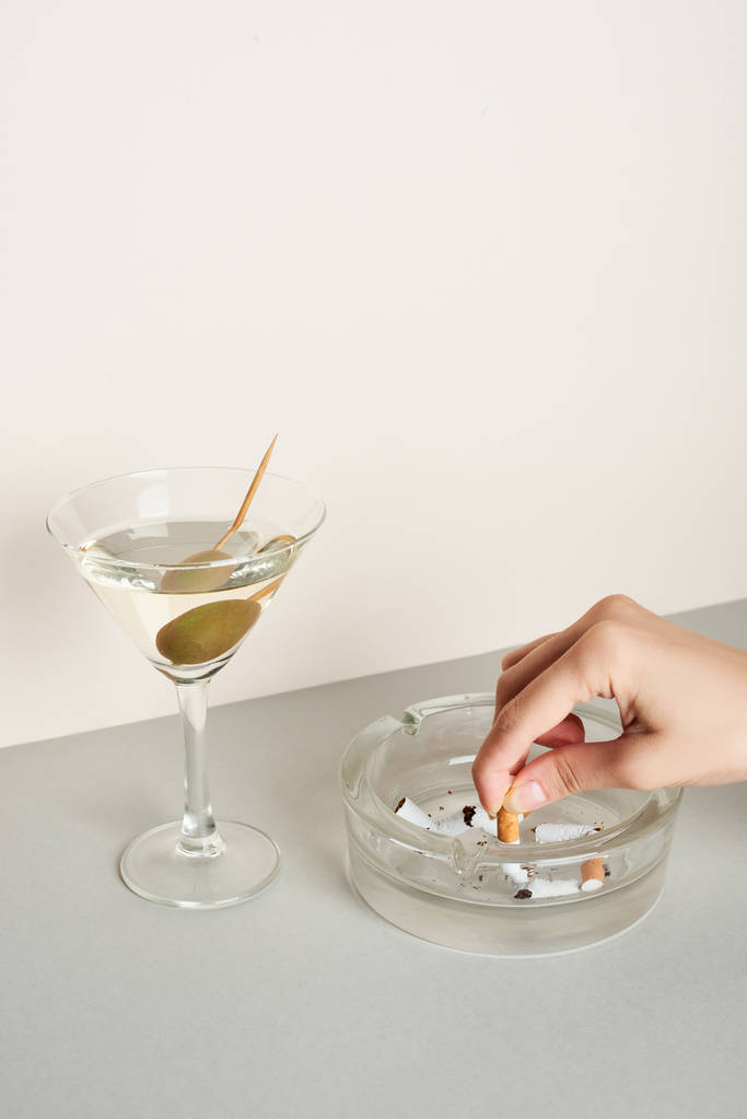 Vista recortada de la mujer poniendo cigarrillo al cenicero con cóctel de martini en la superficie gris
 - Foto, Imagen