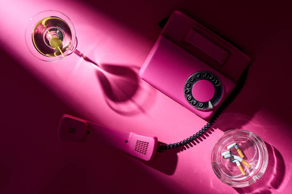 Top view of martini, pink telephone and astray with cigarette butts on pink surface - Photo, Image