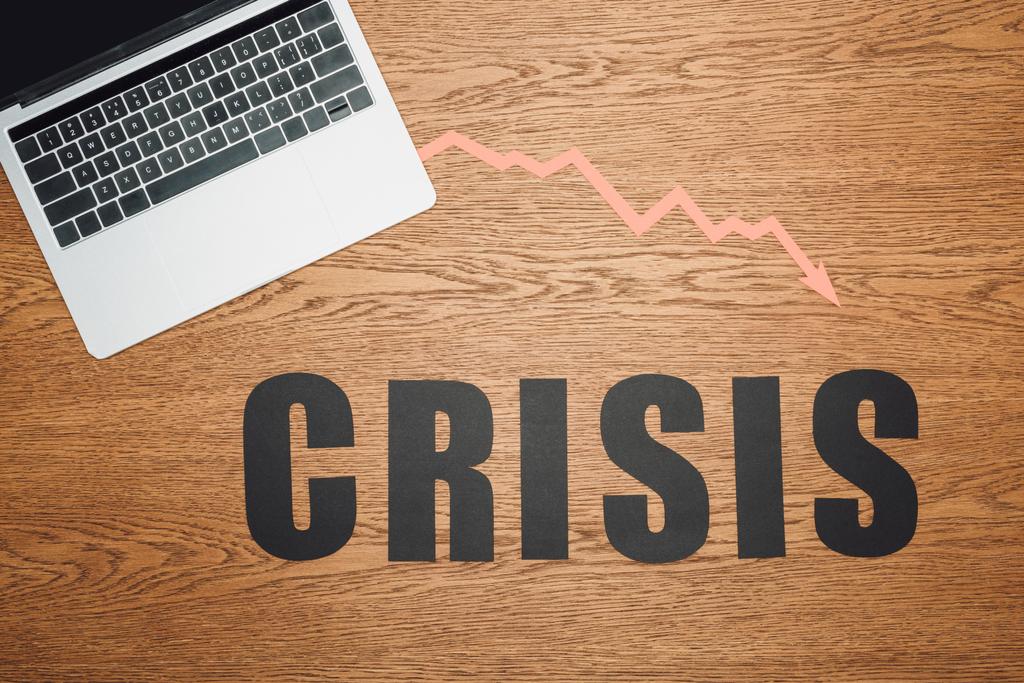 bovenaanzicht van zwart papier gesneden woord crisis in de buurt van laptop en recessie pijl op houten oppervlak - Foto, afbeelding
