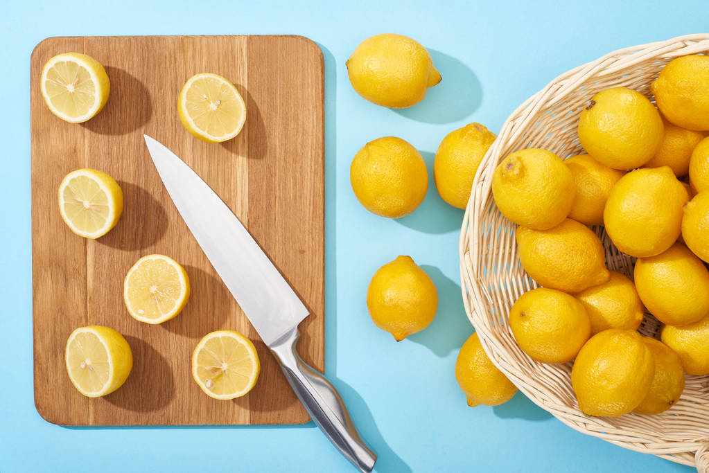 bovenaanzicht van rijpe gele gesneden citroenen op houten snijplank met mes op blauwe achtergrond bij hele citroenen in rieten mand - Foto, afbeelding