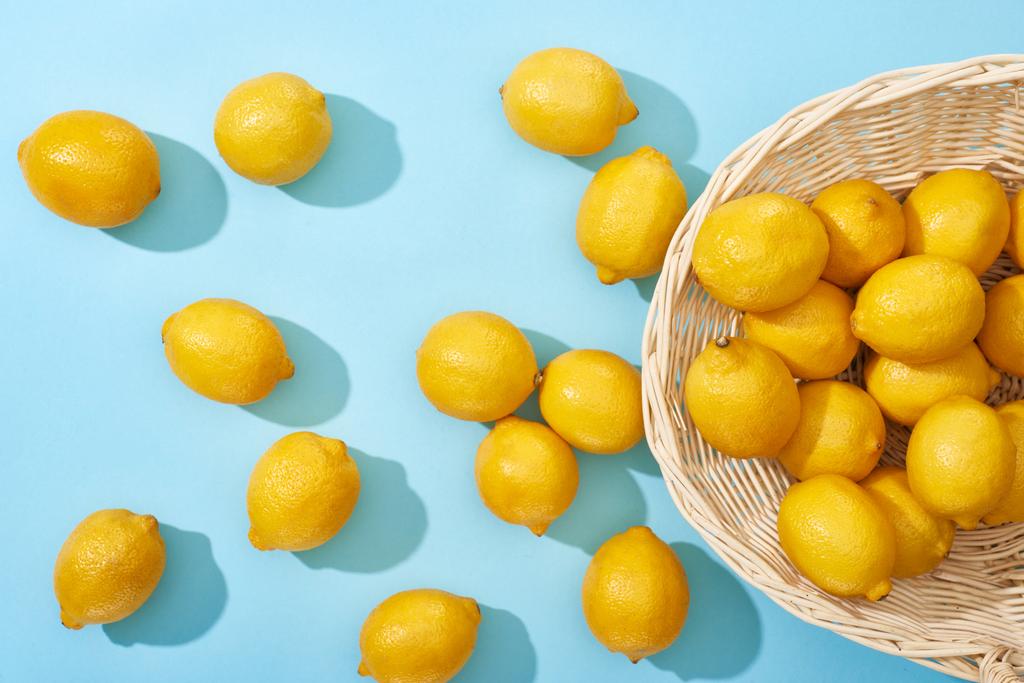 bovenaanzicht van rijpe gele citroenen verspreid uit rieten mand op blauwe achtergrond - Foto, afbeelding