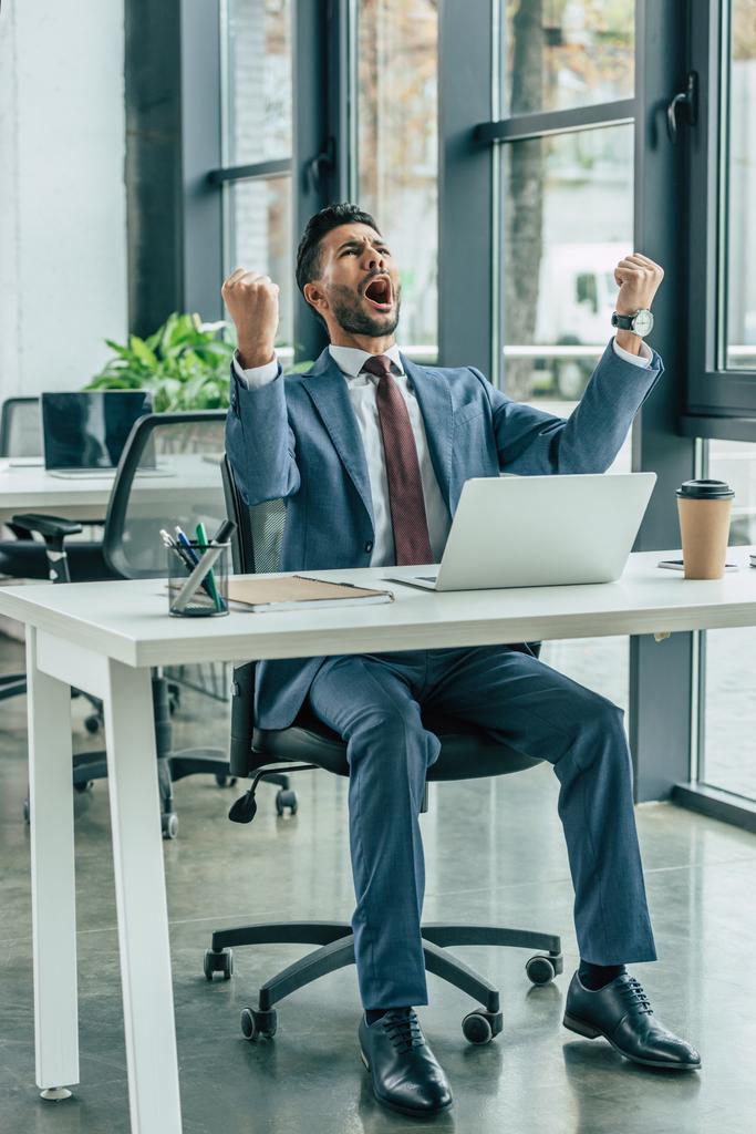 šťastný podnikatel křičí a ukazuje vítězný gesto při sezení na pracovišti - Fotografie, Obrázek