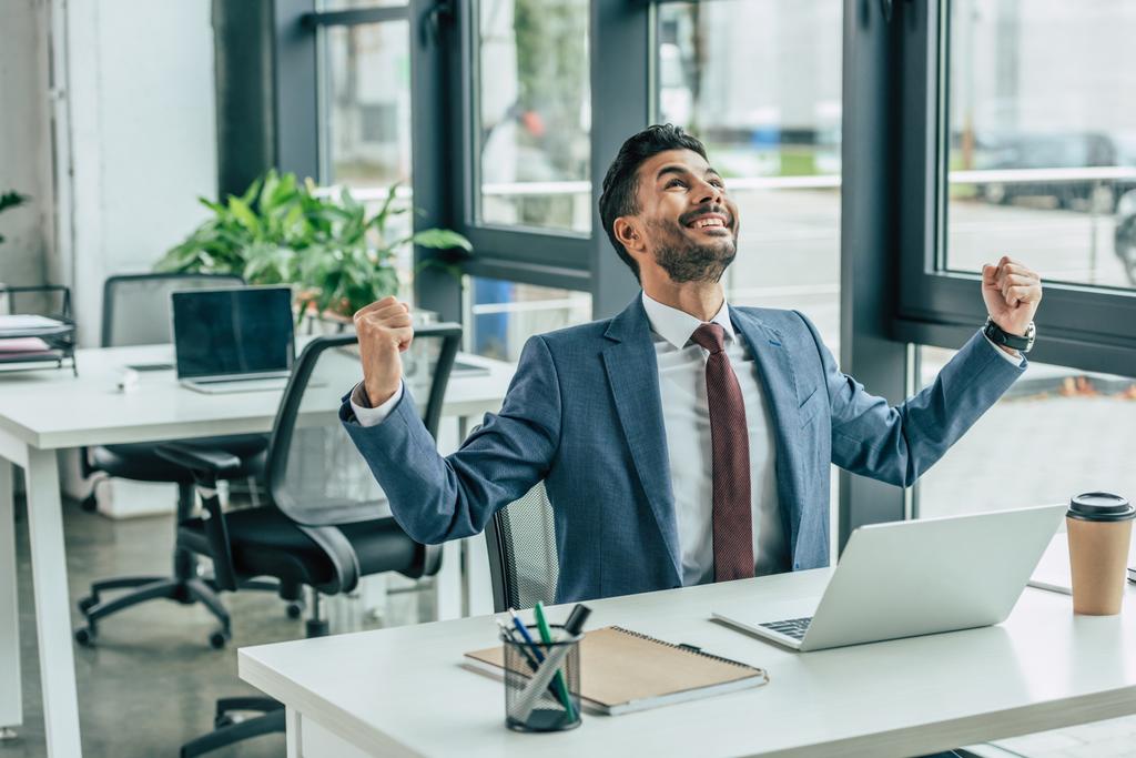 χαρούμενος επιχειρηματίας κοιτάζει ψηλά και δείχνει το νικητή χειρονομία, ενώ κάθεται στο χώρο εργασίας - Φωτογραφία, εικόνα