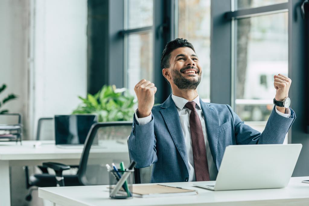 šťastný podnikatel vzhlíží a ukazuje vítězný gesto při sezení na pracovišti - Fotografie, Obrázek