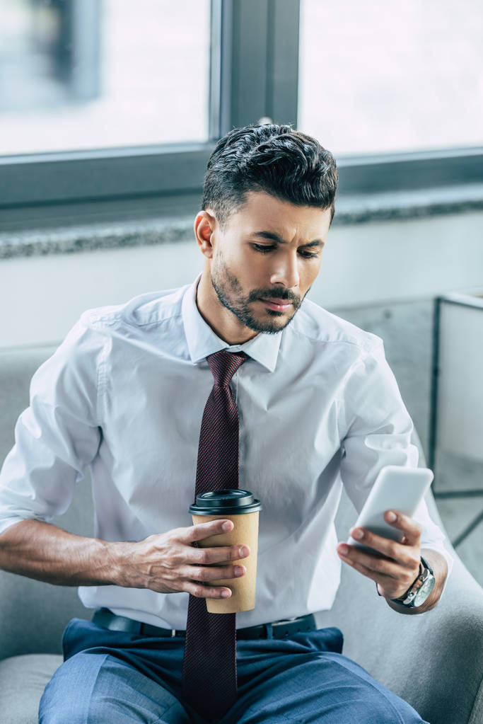 серьезный бизнесмен сидит в кресле, используя смартфон и держа кофе, чтобы пойти
 - Фото, изображение