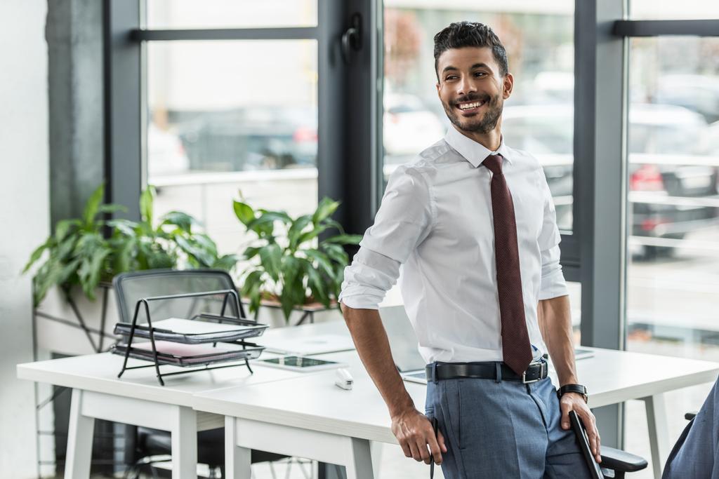νέος επιχειρηματίας χαμογελώντας και κοιτάζοντας μακριά, ενώ στέκεται στο χώρο εργασίας - Φωτογραφία, εικόνα