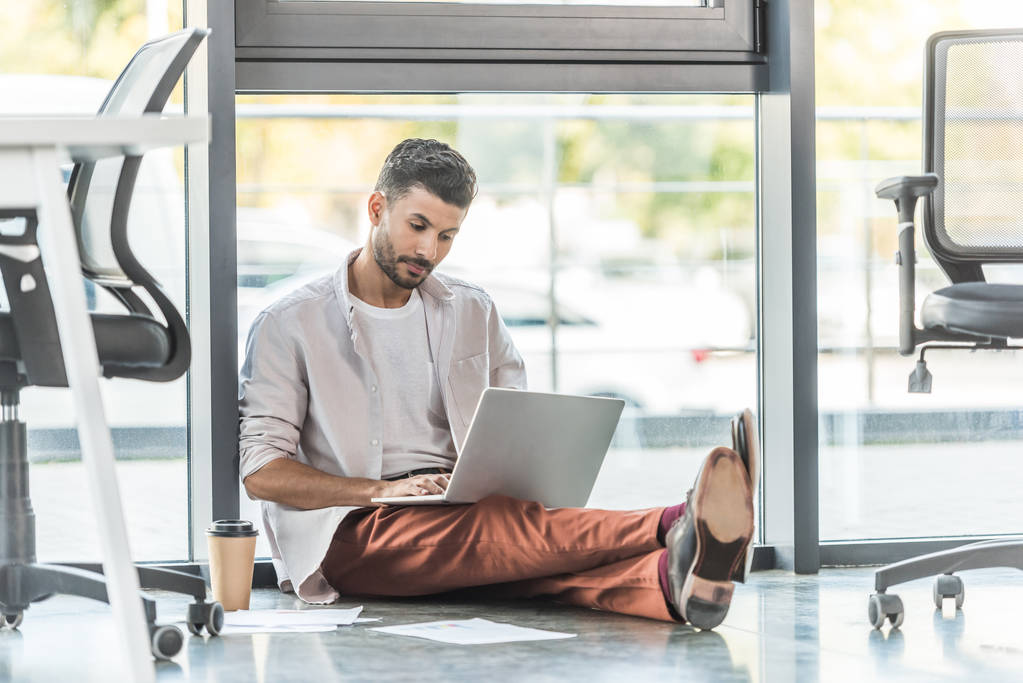 homem de negócios concentrado em roupas casuais sentado no chão perto da janela e usando laptop
 - Foto, Imagem