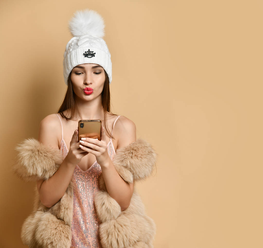 junge Frau im Partykleid, Pelze, die per Handy einen Kuss schicken, Gespräche per Videoanruf, Gruß auf beige mit Kopierplatz - Foto, Bild