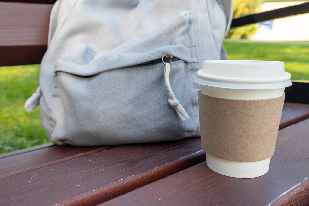 Ένα χάρτινο φλιτζάνι καφέ μιας χρήσης και ένα σακίδιο σε ένα παγκάκι στο πάρκο, φρένο καφέ στην ύπαιθρο - Φωτογραφία, εικόνα