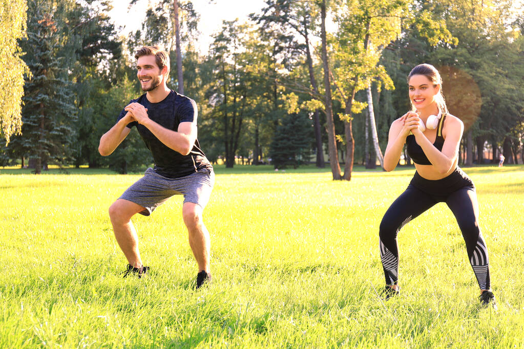 Young sporty man and woman doing workout and squatting together in green park during summer sunny day. - Photo, Image