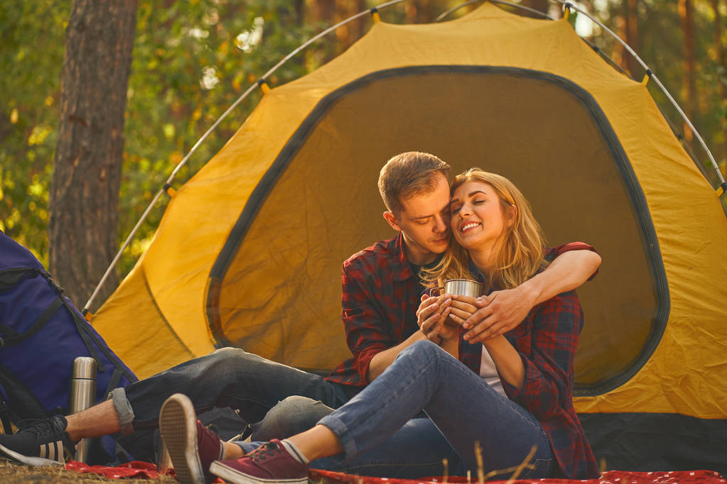 Ormanda kamp yapan bir çift. Çift, ormanın arka planında çadırın yanındaki metal bardaktan kahve içiyor.. - Fotoğraf, Görsel