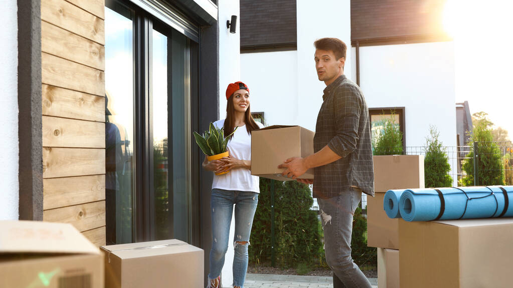 Happy couple with moving boxes and household stuff near their new house on sunny day - 写真・画像