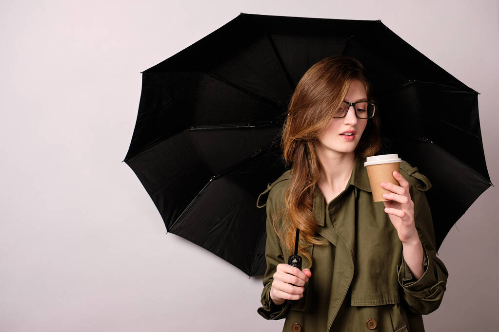 Una chica con una gabardina y gafas de color verde oscuro está bajo un paraguas negro y sostiene una taza de papel en su mano.
.  - Foto, Imagen