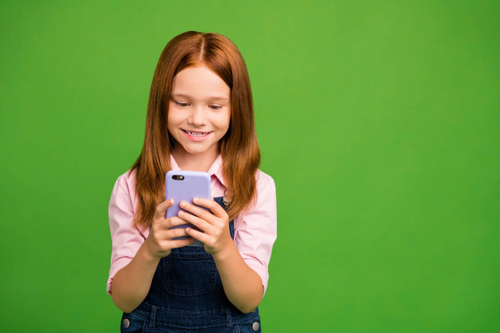 Photo of pretty little schoolchild holding telephone hands texting with classmates friends reading sms wear denim overall pink shirt isolated green background - 写真・画像