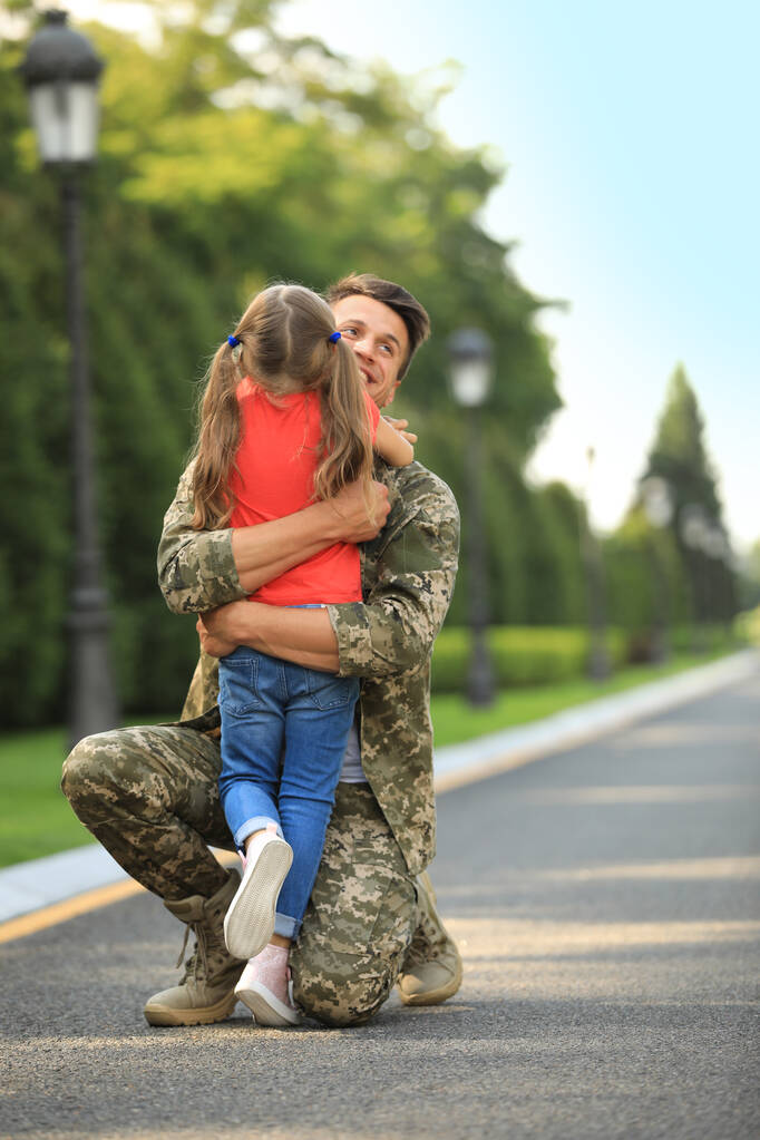 Батько у військовій формі обіймає свою маленьку доньку в сонячному парку
 - Фото, зображення