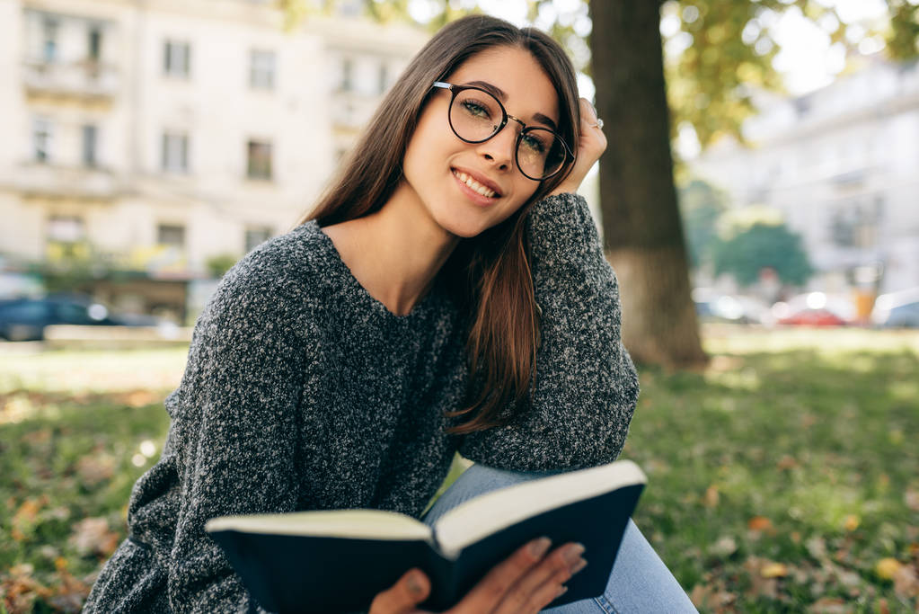 美しい笑みを浮かべて若い女性がセーターや透明な眼鏡を着て、市内公園の屋外に座って、本を読んでのイメージ。街で学ぶ若い女子学生.  - 写真・画像