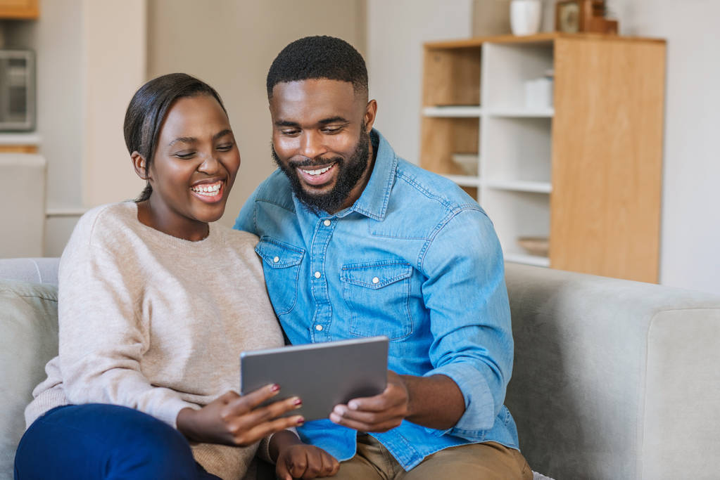 Смеющаяся молодая афроамериканская пара, сидящая вместе на диване в своей гостиной, транслирует видео онлайн с помощью цифрового планшета
 - Фото, изображение