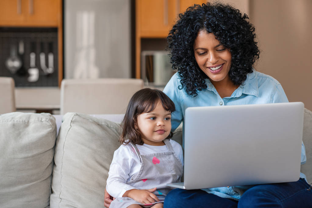 Smiling mother and her cute little daughter watching something together on a laptop while sitting on their living room sofa - Photo, Image