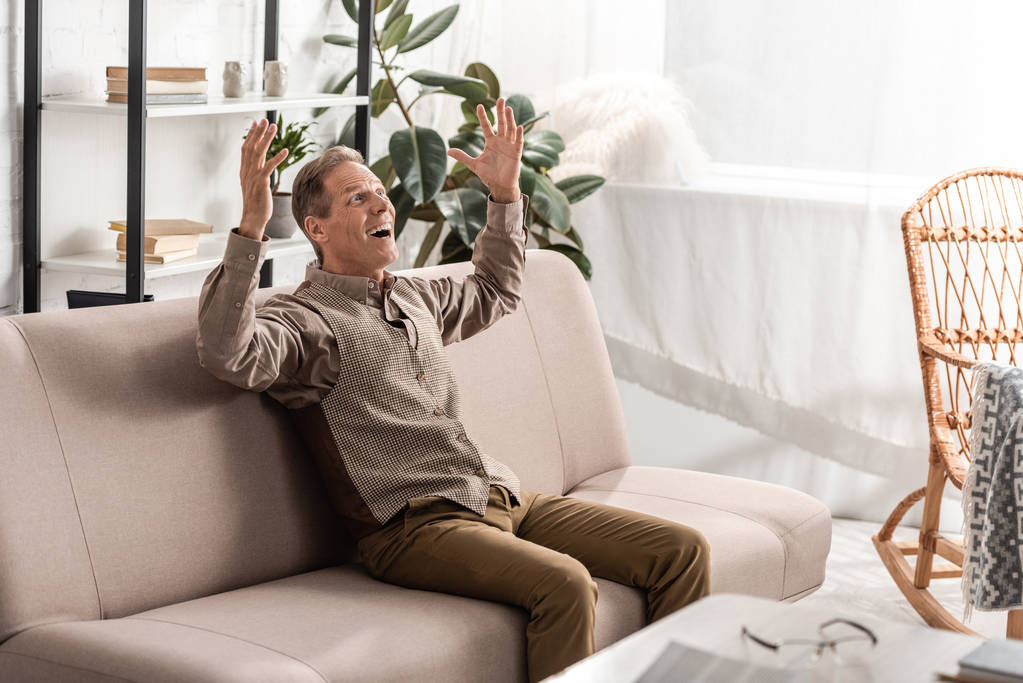 alegre hombre mayor sentado en el sofá y haciendo gestos en casa
  - Foto, Imagen