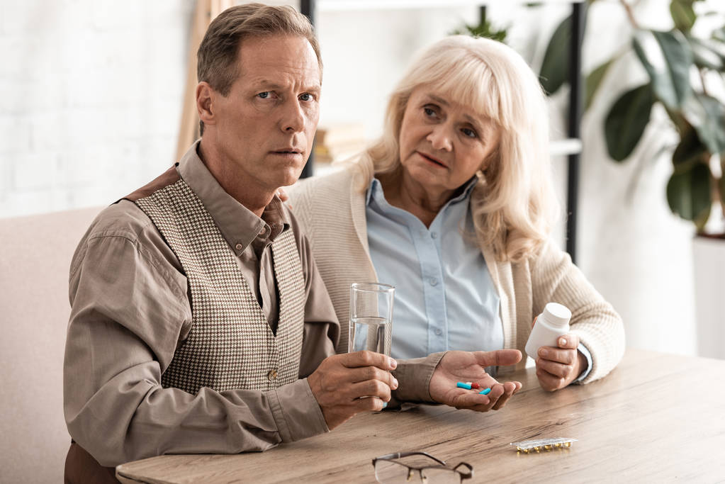 старша жінка сидить з хворим чоловіком тримає склянку з водою і таблетками
  - Фото, зображення