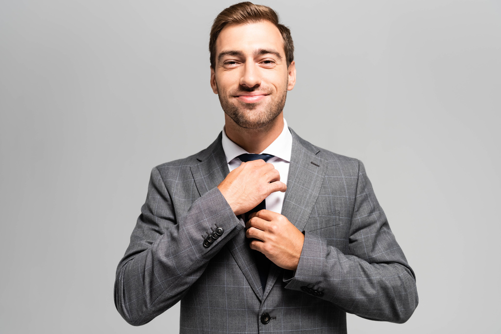 pohledný a usměvavý podnikatel v obleku dotýkající se kravaty izolované na šedé  - Fotografie, Obrázek