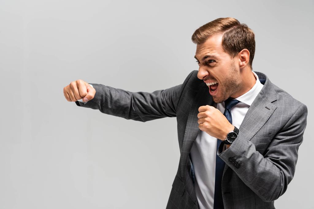 θυμωμένος και όμορφος επιχειρηματίας με κοστούμι χτυπάει απομονωμένος σε γκρι  - Φωτογραφία, εικόνα
