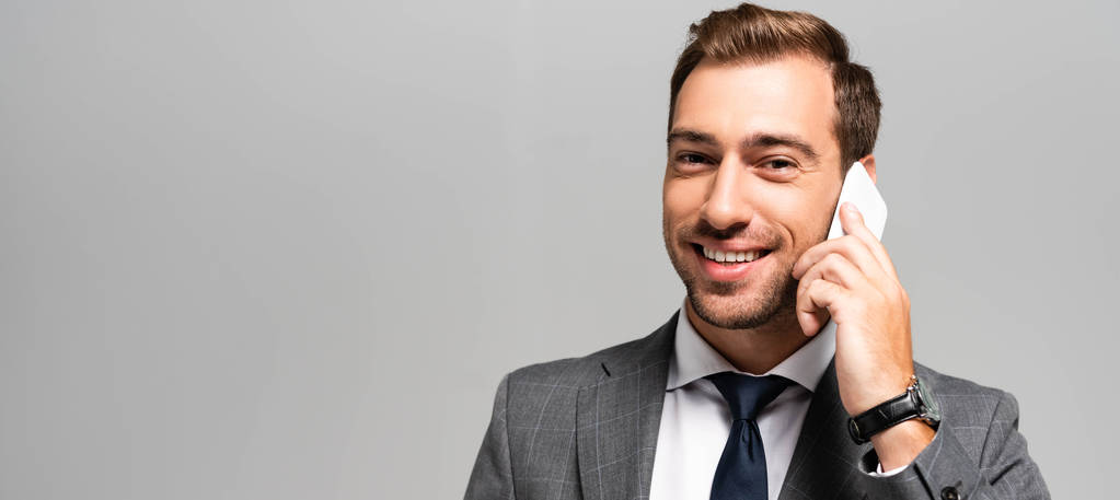 panoramatický záběr pohledný a usmívající se podnikatel v obleku mluví na smartphone izolované na šedé  - Fotografie, Obrázek