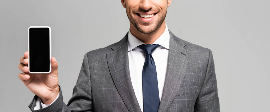Panoramaaufnahme eines lächelnden Geschäftsmannes im Anzug, der sein Smartphone isoliert auf grau hält  - Foto, Bild