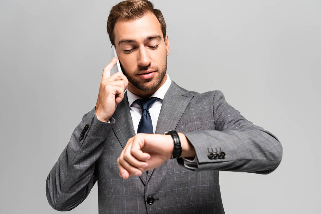 hombre de negocios guapo de traje hablando en el teléfono inteligente y mirando reloj de pulsera aislado en gris
  - Foto, Imagen