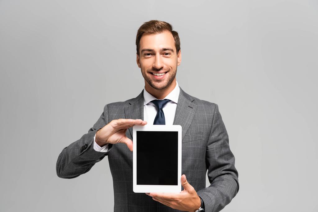 knappe en lachende zakenman in pak met digitale tablet geïsoleerd op grijs  - Foto, afbeelding