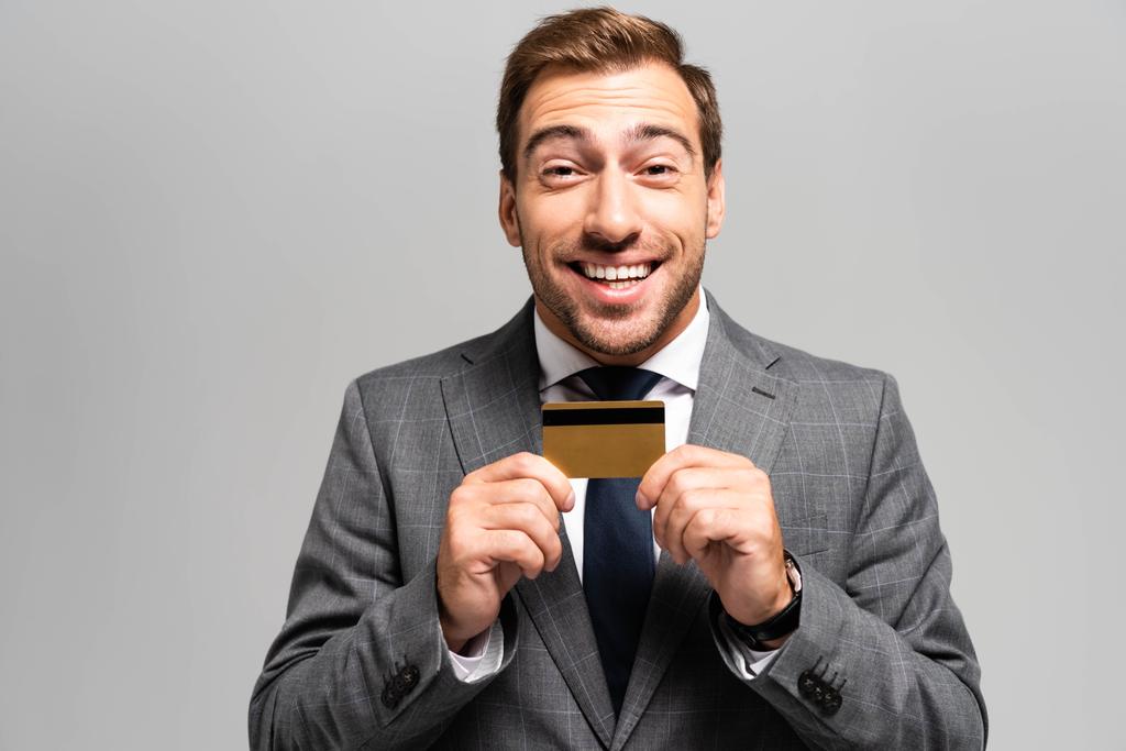 handsome and smiling businessman in suit holding credit card isolated on grey  - Photo, Image