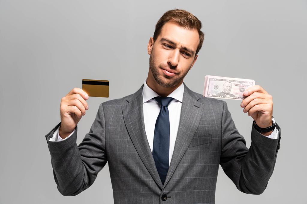 schöner und trauriger Geschäftsmann im Anzug, der Kreditkarten- und Dollarscheine isoliert auf grau hält  - Foto, Bild