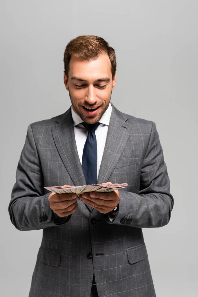 όμορφος και χαμογελαστός επιχειρηματίας με κοστούμι που κρατά τραπεζογραμμάτια δολαρίων απομονωμένα σε γκρι  - Φωτογραφία, εικόνα