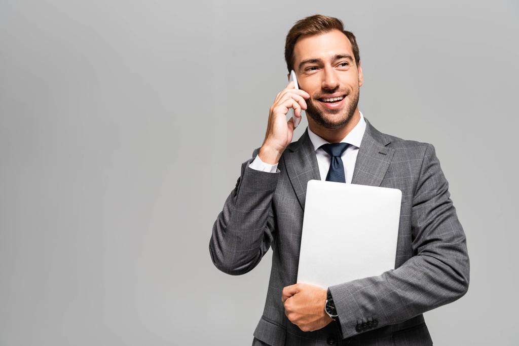 komea ja hymyilevä liikemies puku pitämällä kannettavaa tietokonetta ja puhumalla älypuhelimella eristetty harmaa
  - Valokuva, kuva