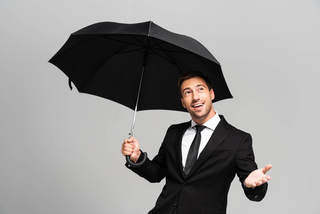 knappe en lachende zakenman met uitgestrekte hand in pak met paraplu geïsoleerd op grijs  - Foto, afbeelding