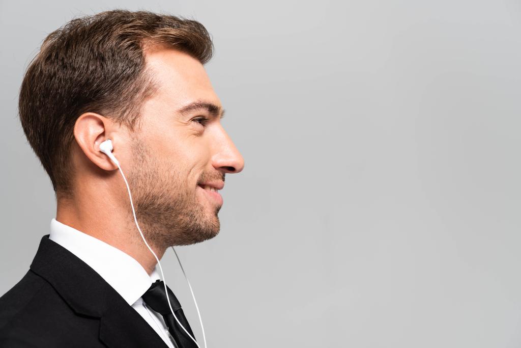 zijaanzicht van knappe en lachende zakenman in pak luistermuziek met koptelefoon geïsoleerd op grijs  - Foto, afbeelding