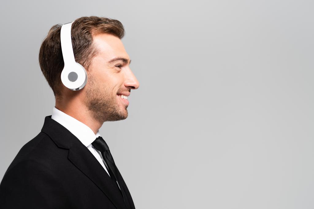 boční pohled na usmívajícího se podnikatele v obleku se sluchátky poslech hudby izolované na šedé  - Fotografie, Obrázek