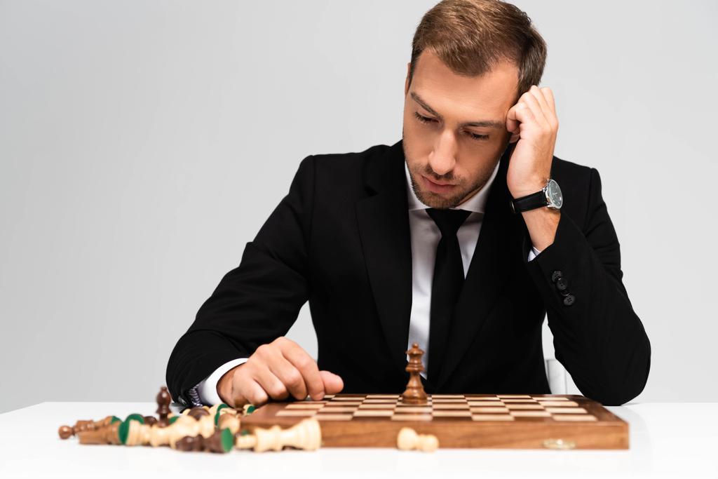 Trauriger und gutaussehender Geschäftsmann im Anzug blickt auf ein Schachbrett isoliert auf grau  - Foto, Bild