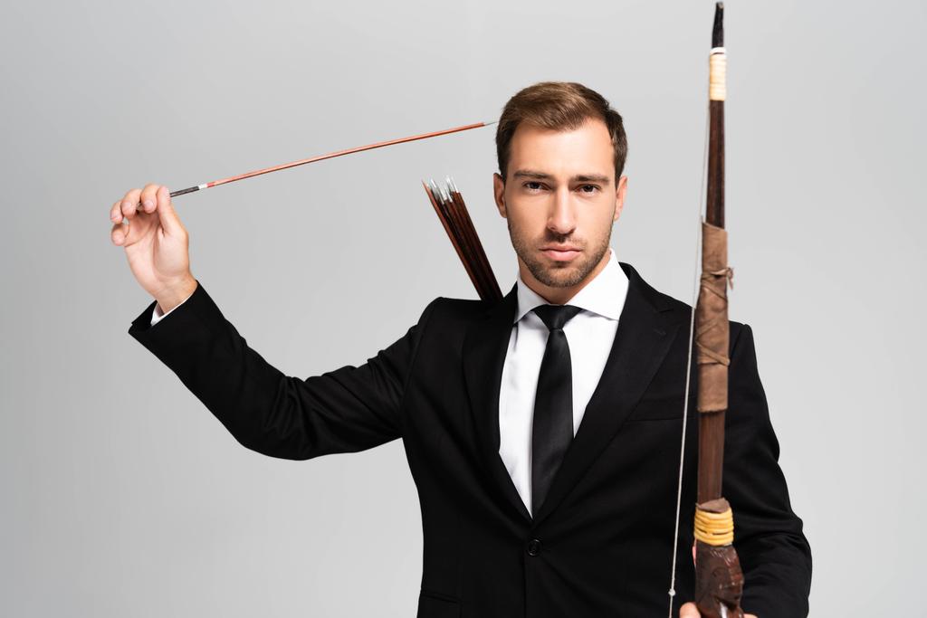 handsome businessman in suit holding bow and arrow isolated on grey  - Photo, Image