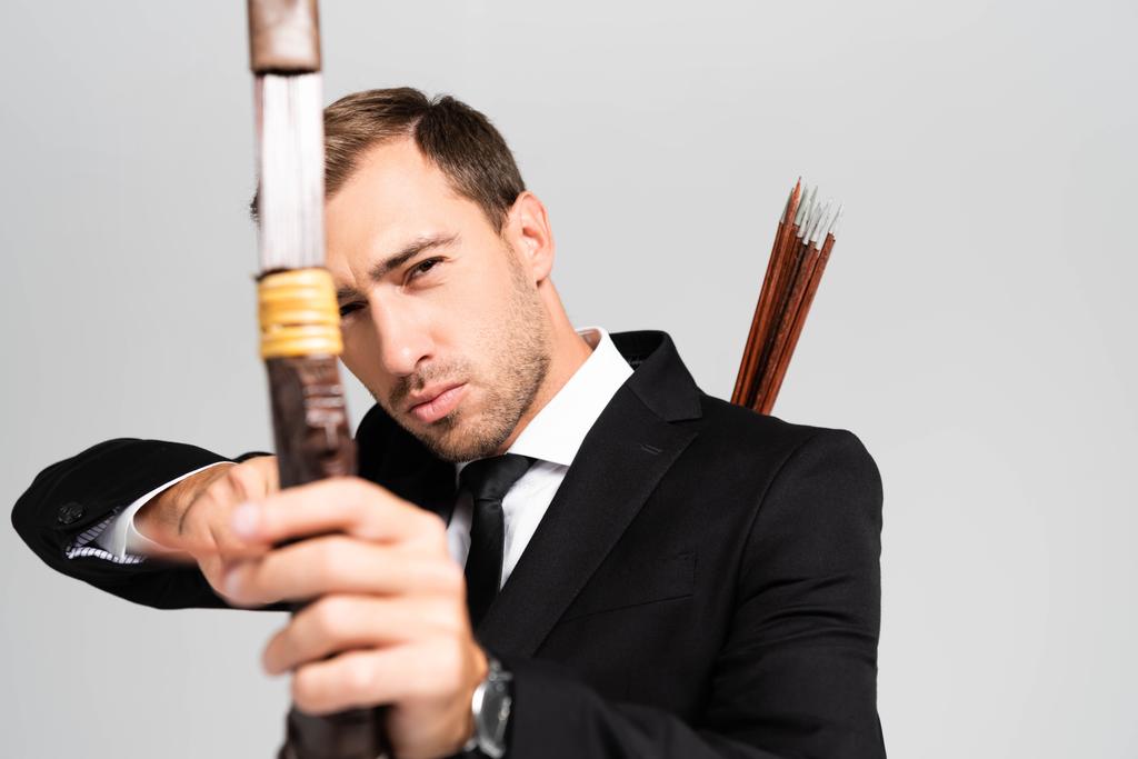 selective focus of handsome businessman in suit holding bow and shooting with arrow isolated on grey - Photo, Image