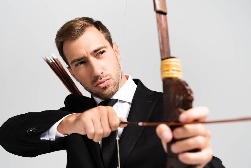 селективное внимание привлекательного бизнесмена в костюме, держащего лук и стреляющего стрелами, изолированными на сером
 - Фото, изображение