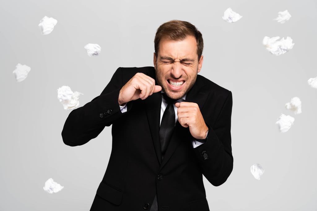 handsome and scared businessman in suit standing near falling crumpled papers isolated on grey - 写真・画像